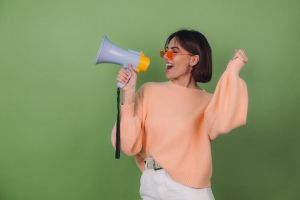 Young woman in casual peach and orange eyeglasses sweater isolated on green olive background happy screaming in megaphone copy space